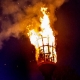 A large beacon fire burns atop a tall metal basket at night, with a crowd of people and trees silhouetted in the background.