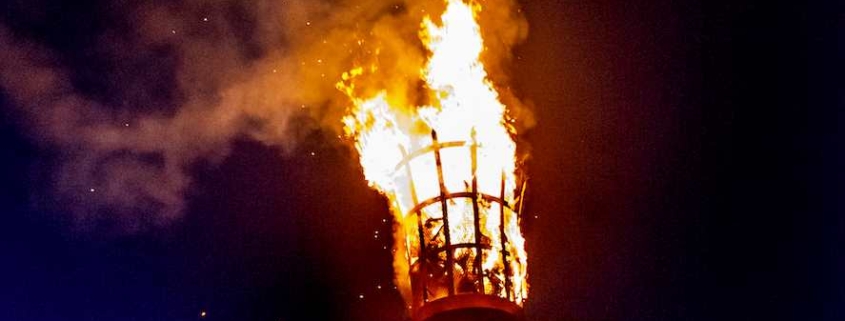 A large beacon fire burns atop a tall metal basket at night, with a crowd of people and trees silhouetted in the background.