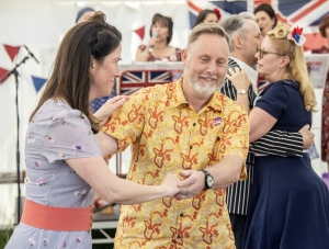People in vintage-style clothing dance together at an indoor event decorated with bunting and Union Jack flags.