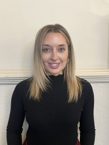 A woman with straight blonde hair wearing a black ribbed sweater stands against a light-colored wall and smiles at the camera.