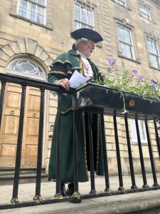 A man dressed in a traditional green ceremonial outfit and tricorn hat stands behind a railing, holding papers, in front of a historic stone building.