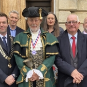 A group of eight adults poses for a photo outside a stone building; the man in the center wears ceremonial green attire and a tricorn hat.