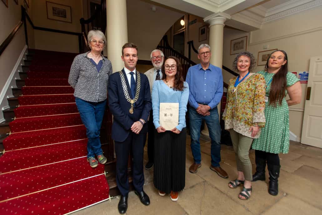 Seven people stand indoors at the base of a red-carpeted staircase; one person holds a certificate, and another wears a ceremonial chain.