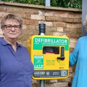 Three adults stand next to a defibrillator station and a parking meter in a car park, with a brick wall and a sign for South Kesteven District Council in the background.