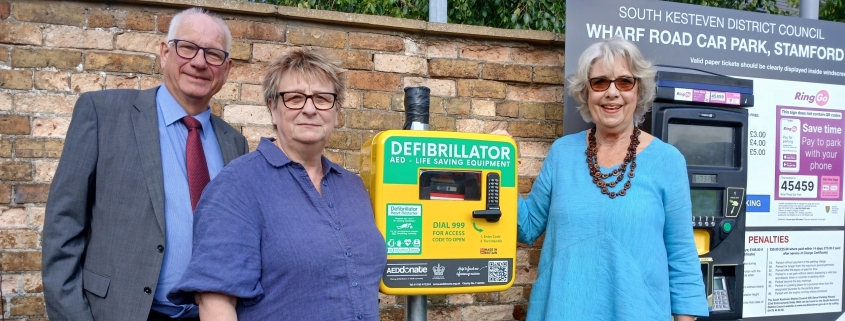 Three adults stand next to a defibrillator station and a parking meter in a car park, with a brick wall and a sign for South Kesteven District Council in the background.