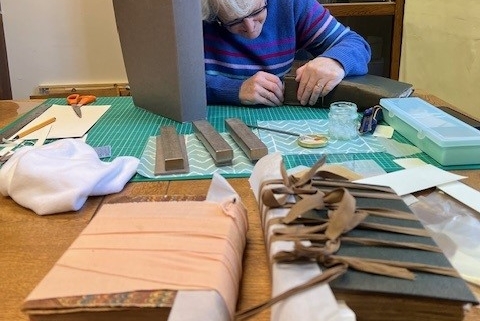 A volunteer works at a table covered with old books, tools, paper, and supplies, with a bookshelf in the background.