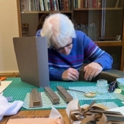 A volunteer works at a table covered with old books, tools, paper, and supplies, with a bookshelf in the background.