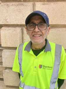 A man wearing glasses, a blue cap, and a high-visibility "Stamford Town Council" vest stands in front of a beige brick wall.
