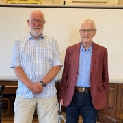 Two men stand side by side indoors in front of a white projection screen. One wears a checked shirt and shorts, the other a red blazer and blue jeans.