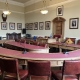 A council chamber with a U-shaped desk, red leather chairs, desktop microphones, framed portraits on the walls, and a wooden door at the back.