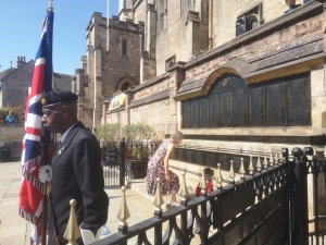 A man in uniform holds a British flag near a memorial wall, while a woman in a floral dress examines names inscribed on the wall outside a historic stone building.
