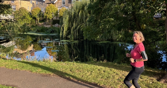 A woman jogs on a paved path beside a pond, with trees and buildings reflected in the water under a clear blue sky.