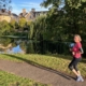 A woman jogs on a paved path beside a pond, with trees and buildings reflected in the water under a clear blue sky.