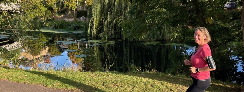 A woman jogs on a paved path beside a pond, with trees and buildings reflected in the water under a clear blue sky.