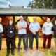 Seven people standing in a row outside a wooden building with warm lighting inside, posing for a group photo in casual and work attire.