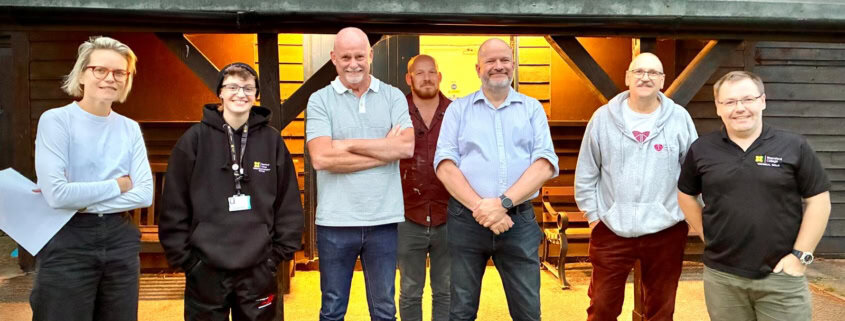 Seven people standing in a row outside a wooden building with warm lighting inside, posing for a group photo in casual and work attire.
