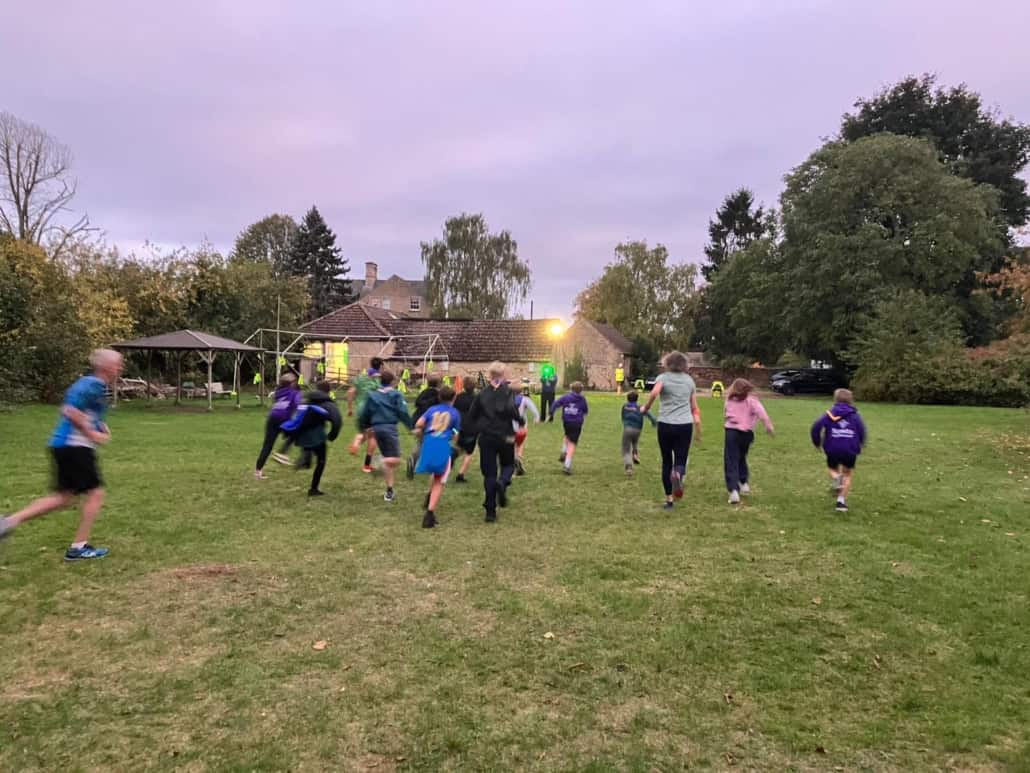 A group of children run across a grassy field toward a building at dusk, with trees and outdoor structures in the background.