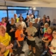 A group of smiling adults in sportswear gather indoors, some holding drinks and food, appearing to celebrate after a running event.