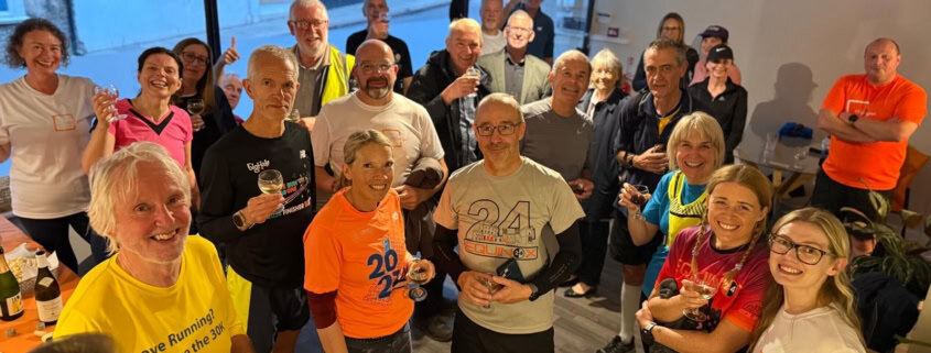A group of smiling adults in sportswear gather indoors, some holding drinks and food, appearing to celebrate after a running event.
