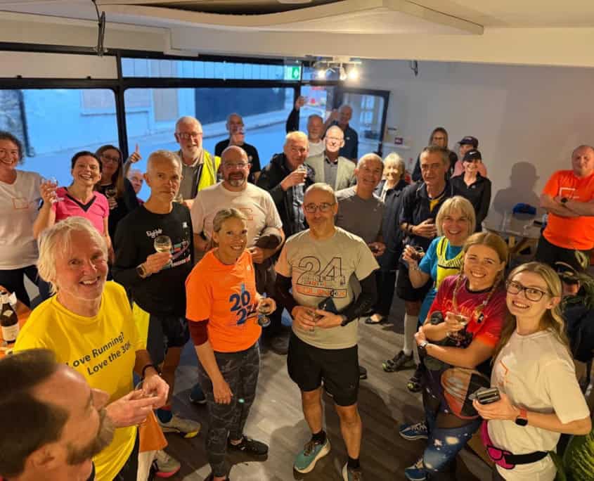A group of smiling adults in sportswear gather indoors, some holding drinks and food, appearing to celebrate after a running event.