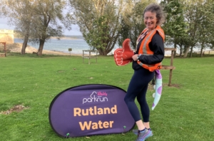 A woman in an orange safety vest stands beside a "parkrun Rutland Water" sign, holding a large foam hand, with trees and a lake in the background.