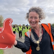 A smiling woman wearing a ceremonial chain and orange vest holds a large red foam thumbs-up sign on a park path, with volunteers in yellow vests in the background.