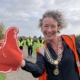 A smiling woman wearing a ceremonial chain and orange vest holds a large red foam thumbs-up sign on a park path, with volunteers in yellow vests in the background.