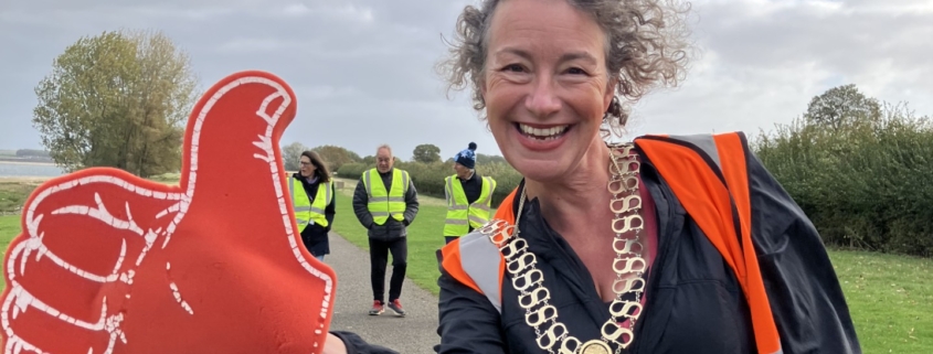 A smiling woman wearing a ceremonial chain and orange vest holds a large red foam thumbs-up sign on a park path, with volunteers in yellow vests in the background.