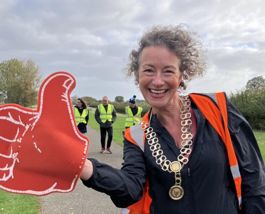 A smiling woman wearing a ceremonial chain and orange vest holds a large red foam thumbs-up sign on a park path, with volunteers in yellow vests in the background.