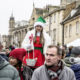 A woman in a Christmas costume and green hat stands above a crowd at an outdoor event, holding a basket with candy canes.