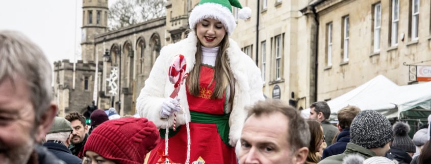 A woman in a Christmas costume and green hat stands above a crowd at an outdoor event, holding a basket with candy canes.