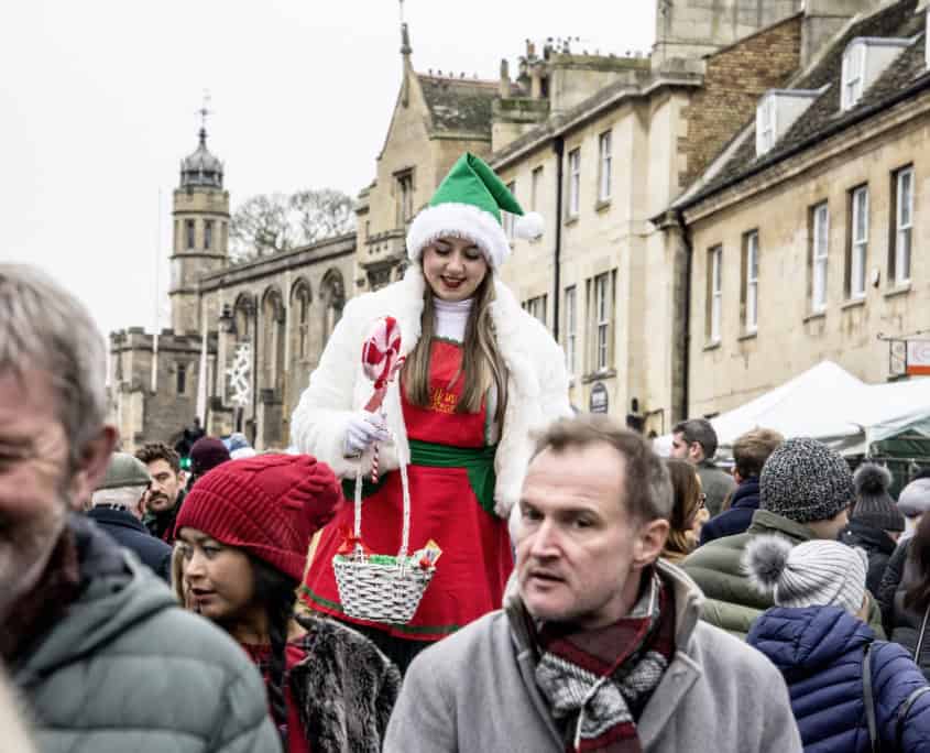 A woman in a Christmas costume and green hat stands above a crowd at an outdoor event, holding a basket with candy canes.