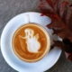 white ceramic teacup filled with ghost illustration coffee latte on white ceramic saucer beside maroon leaf photography
