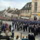 A large military parade with soldiers in uniform, veterans, and officials standing in formation on a street lined with historic buildings and spectators.