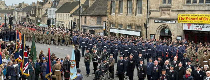 A large military parade with soldiers in uniform, veterans, and officials standing in formation on a street lined with historic buildings and spectators.