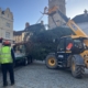 A yellow JCB telehandler is lifting a large fir tree onto a flatbed truck in a cobbled town square, as a worker looks on. A historic church is visible in the background.