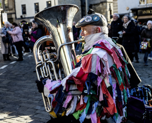 An older man wearing a colorful patchwork outfit plays a large brass tuba on a cobblestone street, surrounded by people in an outdoor setting.