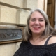 A woman with gray hair and a black top stands smiling in front of a stone building with a sign reading "TOWN HALL.