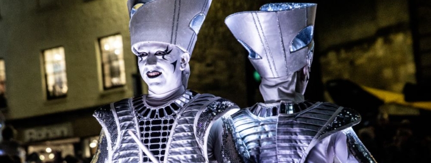 Two performers in futuristic metallic costumes and makeup play illuminated drums at night during an outdoor event.