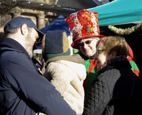 Four people, including a child in winter clothes, gather outdoors. One adult wears a festive red hat and costume, smiling at the group under a blue canopy.