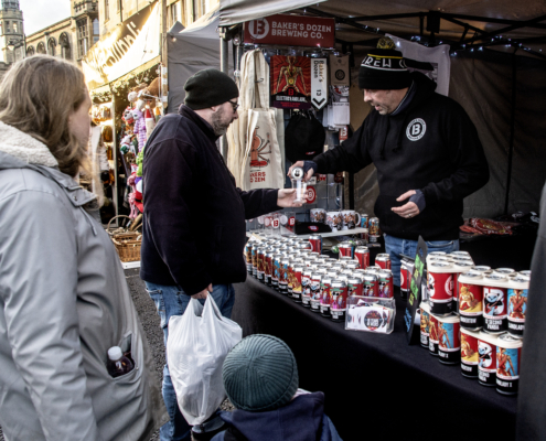 A vendor at a market stall hands a canned drink to a customer; various cans are displayed on the table while other people stand nearby.