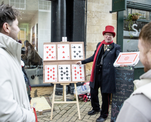 A street performer in a red hat and scarf displays large playing cards on a board to an audience during a card trick on a city sidewalk.