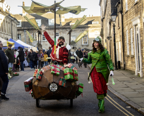 Two people in festive, aviator-themed costumes push a propellered cart decorated with wrapped presents during a street festival with onlookers and market stalls.