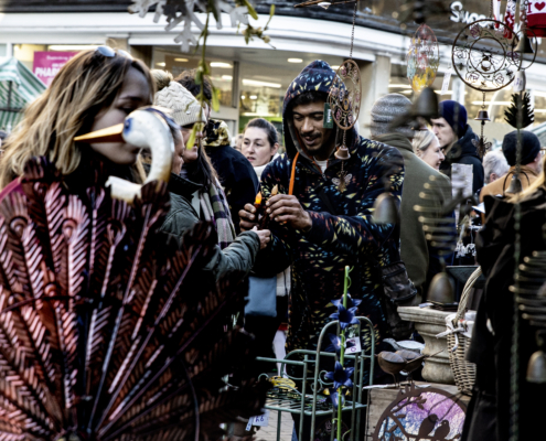 A busy outdoor market scene with people browsing various crafts and decorations; a man in a patterned hoodie examines an item while others look on.