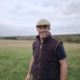 A man wearing a flat cap, dark vest, and t-shirt stands smiling in a grassy field with a cloudy sky in the background.