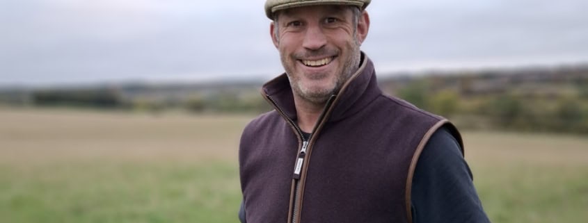 A man wearing a flat cap, dark vest, and t-shirt stands smiling in a grassy field with a cloudy sky in the background.
