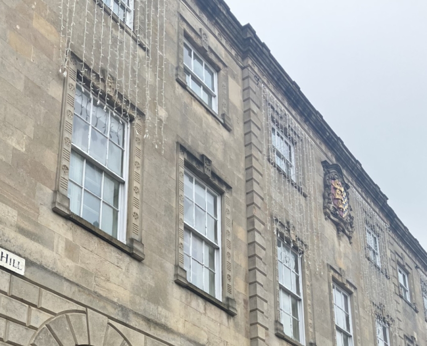 Three-story stone building with arched windows, a British flag on a pole, a crest above the entrance, and a potted plant by a black railing. Street sign reads "St Mary's Hill.