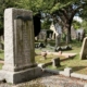 A stone grave marker stands in a cemetery surrounded by grass, trees, and several other headstones.