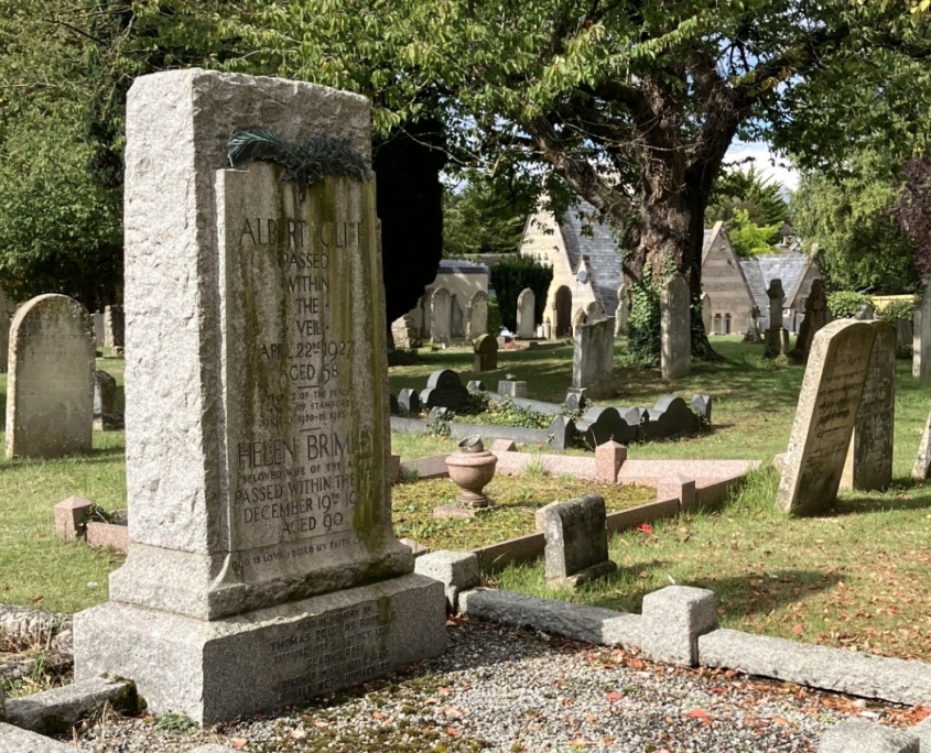 A stone grave marker stands in a cemetery surrounded by grass, trees, and several other headstones.
