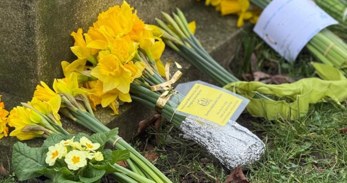 Several bouquets of yellow flowers and a small potted plant are placed at the base of a stone monument on grass.